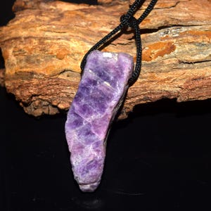 Puede incluir: Un colgante de amatista púrpura en bruto cuelga de un cordón negro. La piedra preciosa es de color violeta intenso con estructuras cristalinas visibles. El colgante está suspendido sobre un fondo de madera marrón texturizada.
