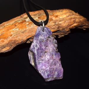 May include: A raw, purple amethyst pendant hangs from a black cord. The irregularly shaped stone has a rough, textured surface with flecks of darker color. The pendant is attached to a silver-colored bail.