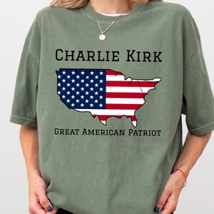 May include: Olive green t-shirt featuring the text "CHARLIE KIRK" above a graphic of the United States flag. The flag design is split, with the stars and stripes representing the American flag. Below the graphic, the text reads "GREAT AMERICAN PATRIOT".