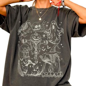 May include: Dark gray t-shirt featuring a Halloween-themed graphic. The design includes a witch, ghosts, skulls, a crow, bats, and a jack-o'-lantern. The shirt has short sleeves and a relaxed fit, perfect for the spooky season.
