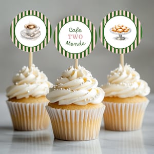 May include: Three cupcakes topped with round paper toppers on wooden picks. The toppers feature green and pink vertical stripes. One shows a coffee cup, one says Cafe TWO Monde, and one shows a plate of pastries. The cupcakes have white frosting.