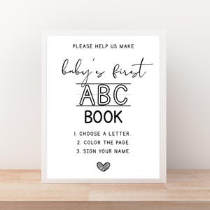 May include: A framed sign with the text "PLEASE HELP US MAKE baby's first ABC BOOK". Instructions include choosing a letter, coloring the page, and signing your name. A heart graphic is at the bottom. The sign is white with black text.