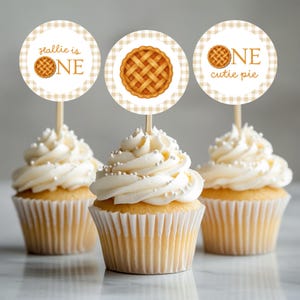 May include: Three cupcakes topped with round paper toppers on wooden sticks. The toppers feature a beige gingham border and illustrations of lattice-crust pies. Text includes Hallie is ONE, a pie graphic, and ONE cutie pie in brown font.