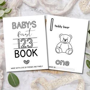 May include: Two white baby books with black text. One book reads "BABY'S first 123 BOOK" with a heart. The other book has a teddy bear illustration and the word "one". Both books are for baby's first learning.
