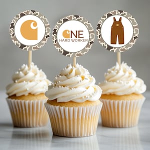 Puede incluir: Tres cupcakes con adornos de papel redondos sobre palitos de madera. Los adornos tienen un borde de camuflaje marrón y blanco. Uno muestra un logo amarillo, otro dice ONE HARD WORKER con un logo, y el tercero muestra un overol marrón con un logo.