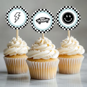 May include: Three cupcakes with white frosting and silver sprinkles. Each cupcake has a decorative topper with a black and white checkered border. The toppers feature a lightning bolt, a skateboard with "Baby on Board" text, and a smiley face.