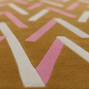 May include: Close-up of a textured rug with a geometric pattern. The rug features a mustard yellow background with white and pink angled lines creating a modern design. The rug's texture is visible.