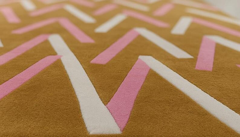May include: Close-up of a textured rug with a geometric pattern. The rug features a mustard yellow background with white and pink angled lines creating a modern design. The rug's texture is visible.
