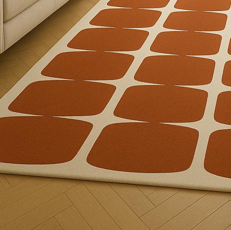 May include: A rectangular rug featuring a repeating pattern of rust-coloured, rounded rectangles against a cream background. The rug is placed on a wooden floor, with a glimpse of a cabinet in the upper left corner.