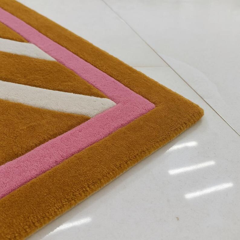 May include: A close-up of a geometric rug with a repeating pattern. The rug features a mustard yellow background with white and pink stripes. The rug's border is a darker shade of mustard yellow.