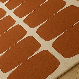 May include: A close-up of a rug featuring a repeating pattern of rust-coloured rectangular shapes with white, pointed extensions. The rug has a soft, textured appearance, and the design is modern and geometric.