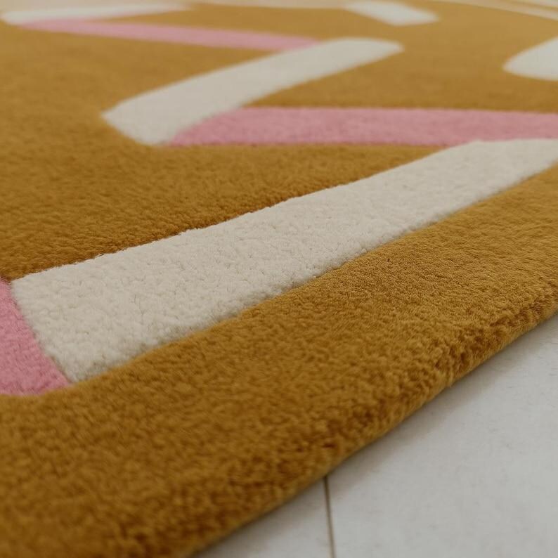 May include: Close-up of a mustard-colored rug with a geometric design. The rug features white and pink stripes and shapes against the mustard background. The rug has a soft, textured surface.