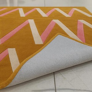 May include: Mustard yellow area rug with a geometric design featuring pink and off-white zig-zag patterns. The rug's underside is visible, revealing a gray, textured backing. The edges are neatly finished with a yellow border.