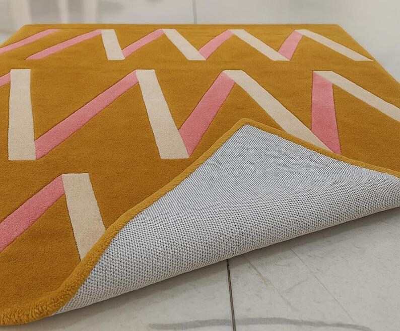 May include: Mustard yellow area rug with a geometric design featuring pink and off-white zig-zag patterns. The rug's underside is visible, revealing a gray, textured backing. The edges are neatly finished with a yellow border.