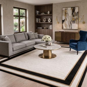 Puede incluir: Una sala de estar con un gran sof&aacute; seccional gris, una mesa de centro redonda de m&aacute;rmol con base de lat&oacute;n y un sill&oacute;n orejero de terciopelo azul intenso. Una alfombra rectangular con centro crema y bordes negros y dorados cubre el suelo.