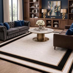 Puede incluir: Una alfombra rectangular grande con un centro de color crema claro, enmarcada por un borde negro grueso y una fina l&iacute;nea dorada. La alfombra est&aacute; situada en una sala de estar con un sof&aacute; gris capiton&eacute; y un sof&aacute; de cuero marr&oacute;n.