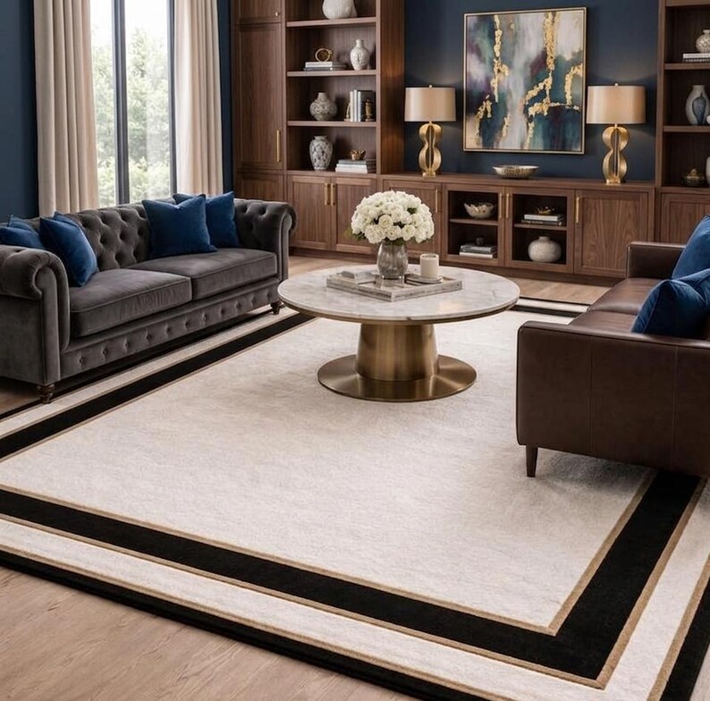Puede incluir: Una alfombra rectangular grande con un centro de color crema claro, enmarcada por un borde negro grueso y una fina l&iacute;nea dorada. La alfombra est&aacute; situada en una sala de estar con un sof&aacute; gris capiton&eacute; y un sof&aacute; de cuero marr&oacute;n.