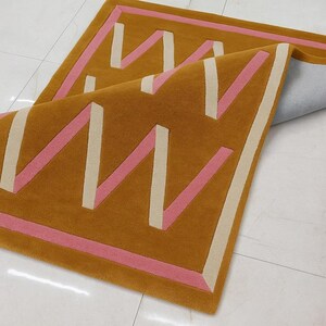 May include: Mustard-colored rectangular rug with a geometric design. The rug features a repeating pattern of pink and off-white zigzags and borders. The rug is partially folded, revealing the backing.