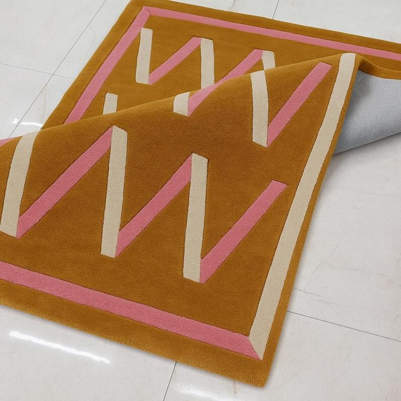 May include: Mustard-colored rectangular rug with a geometric design. The rug features a repeating pattern of pink and off-white zigzags and borders. The rug is partially folded, revealing the backing.