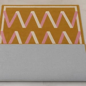 May include: Mustard yellow rug with a geometric zigzag pattern in pink and cream. The rug has a rectangular shape and a gray backing. The design features a repeating series of chevrons, creating a modern and stylish look for home decor.