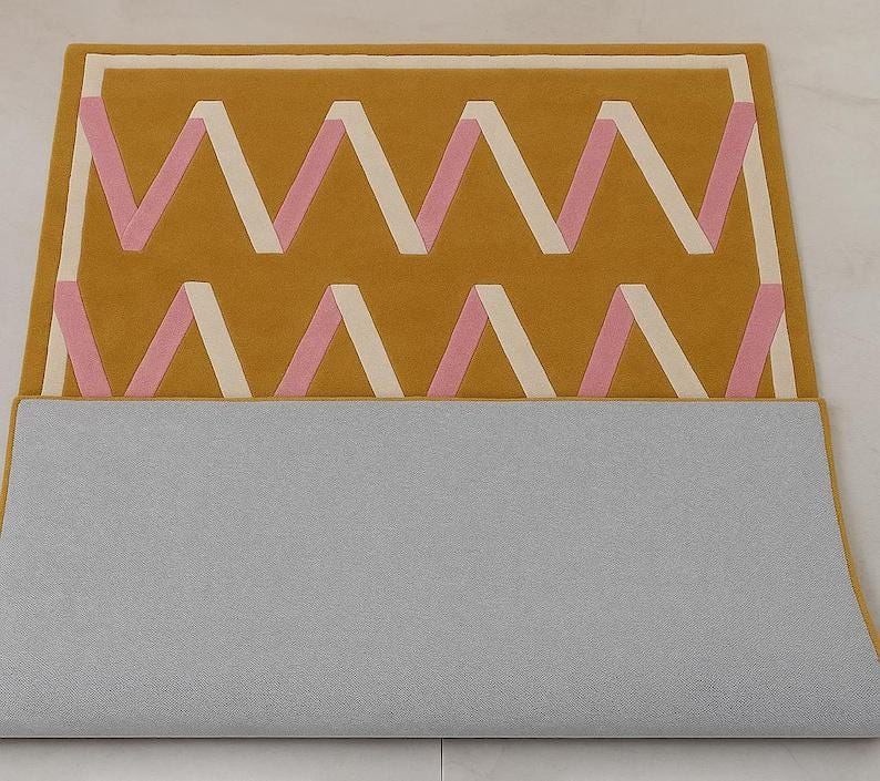 May include: Mustard yellow rug with a geometric zigzag pattern in pink and cream. The rug has a rectangular shape and a gray backing. The design features a repeating series of chevrons, creating a modern and stylish look for home decor.