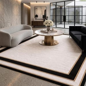 Puede incluir: Una alfombra rectangular grande con un centro color crema, un borde ancho negro y una fina franja dorada. La alfombra est&aacute; colocada sobre un suelo moteado entre un sof&aacute; curvo de color gris claro y un sof&aacute; negro con una mesa de centro.
