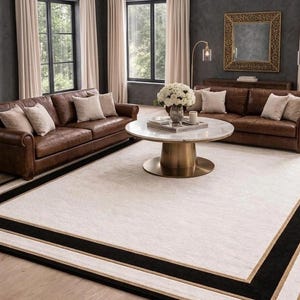 Puede incluir: Una alfombra rectangular grande con un centro de color crema bordeado por una franja negra gruesa y un ribete fino de tono dorado. La alfombra est&aacute; colocada sobre un suelo de madera clara frente a dos sof&aacute;s de cuero marr&oacute;n y una mesa de centro redonda.
