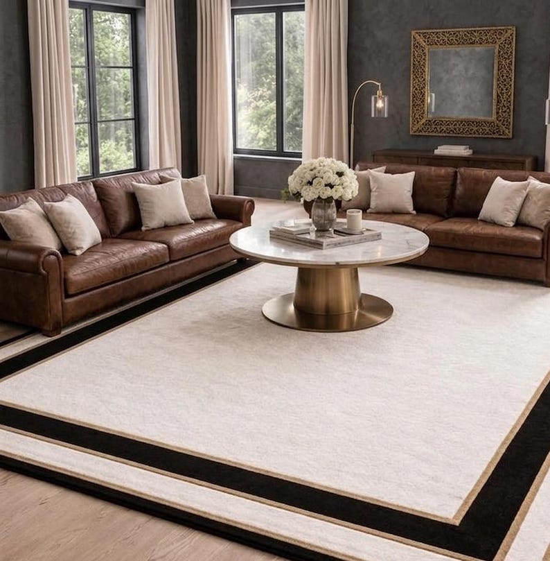 Puede incluir: Una alfombra rectangular grande con un centro de color crema bordeado por una franja negra gruesa y un ribete fino de tono dorado. La alfombra est&aacute; colocada sobre un suelo de madera clara frente a dos sof&aacute;s de cuero marr&oacute;n y una mesa de centro redonda.