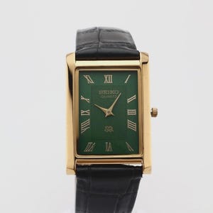 May include: A rectangular Seiko Quartz wristwatch featuring a gold-tone case, a deep green dial with a textured concentric pattern, gold Roman numeral hour markers, gold hands, and a black leather strap with a visible crocodile-style texture.