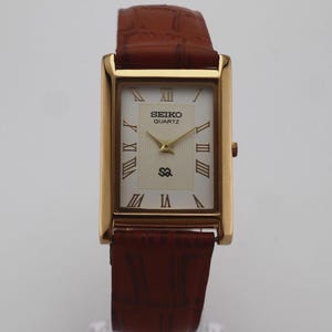 May include: A rectangular Seiko Quartz wristwatch with a gold-tone case and a white dial featuring black Roman numeral hour markers. The watch has gold-tone hands and a brown leather strap with a textured, embossed crocodile-style pattern.
