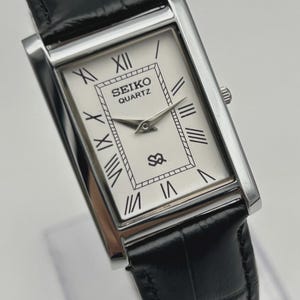 May include: A rectangular Seiko Quartz wristwatch with a silver-tone case and a white dial featuring black Roman numeral hour markers. The watch has silver-tone hands, a small SQ logo, and a black leather strap with a textured crocodile-like pattern.