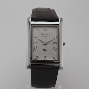Könnte beinhalten: Eine rechteckige Seiko Quartz Armbanduhr mit silberfarbenem Zifferblatt und schwarzen römischen Ziffern. Die Uhr hat ein poliertes silberfarbenes Metallgehäuse und ein dunkelbraunes, strukturiertes Lederarmband mit Krokodilprägung.