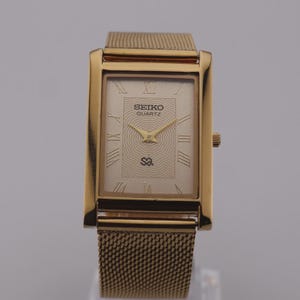 May include: A gold-tone rectangular wristwatch with a mesh strap. The watch face features a light champagne background with a concentric wave pattern, black Roman numeral hour markers, gold hands, and the text SEIKO QUARTZ with a logo.