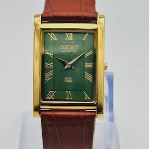 Könnte beinhalten: Eine rechteckige Seiko Quartz Armbanduhr mit goldfarbenem Gehäuse und strukturiertem grünem Zifferblatt mit goldenen römischen Ziffern. Die Uhr verfügt über ein braunes Lederarmband mit Krokodilprägung und goldfarbene Zeiger.