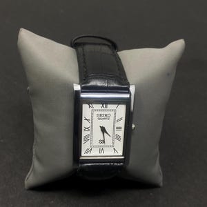 May include: A rectangular Seiko Quartz wristwatch featuring a white dial with black Roman numeral hour markers, black hands, and a silver-tone metal case. The watch is fitted with a black leather strap that has a textured, embossed finish.