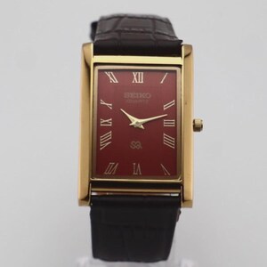 Könnte beinhalten: Eine rechteckige Seiko Quartz Armbanduhr mit goldfarbenem Gehäuse und dunkelrotem Zifferblatt. Das Zifferblatt zeigt goldene römische Ziffern als Stundenmarkierungen, goldene Zeiger und das Seiko-Logo. Sie hat ein dunkelbraunes Lederarmband.