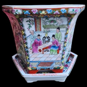 Könnte beinhalten: Ein quadratischer, handbemalter Porzellan-Blumentopf mit passender Untertasse. Der Topf zeigt eine detaillierte Szene von Figuren in traditioneller Kleidung mit floralen und geometrischen Bordüren. Die Farbpalette umfasst rosa, blaue, grüne und goldene Akzente.