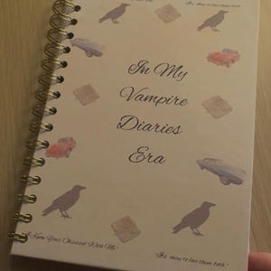 Könnte beinhalten: Ein spiralgebundenes Notizbuch mit cremefarbenem Einband. Der Einband zeigt den Text "In My Vampire Diaries Era" in eleganter Schrift, zusammen mit Illustrationen von Autos, Büchern und Krähen. Das Notizbuch hat eine goldfarbene Spiralbindung.