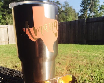 May include: A stainless steel tumbler with a black finish, featuring a peach-colored Texas outline and the name "Amanda" in gold glitter script. Gold-framed aviator sunglasses with orange lenses rest in front of the tumbler.