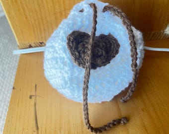 May include: A white crocheted pouch with a brown heart-shaped design in the centre. The pouch has a drawstring closure made of brown yarn. The pouch is on a wooden surface.