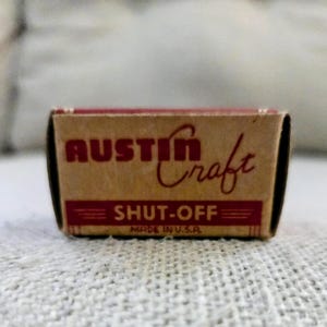 Op de afbeelding: Een kleine rechthoekige kartonnen doos met een beige achtergrond en rode tekst. De voorkant van de doos toont de merknaam AUSTIN Craft in een vetgedrukt lettertype, gevolgd door de tekst SHUT-OFF en de tekst MADE IN U.S.A. onderaan.