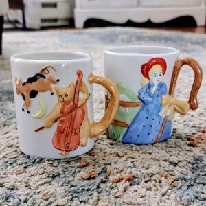 Könnte beinhalten: Zwei weiße Keramiktassen mit skurrilen Designs. Eine Tasse zeigt eine Katze, die Cello spielt, mit einer Ziege und einem Mond. Die andere Tasse zeigt eine Frau in einem blauen Kleid und einem roten Hut mit einem Stab. Beide Tassen haben gebogene Griffe.