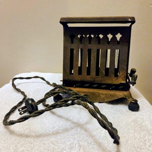 May include: An antique metal toaster with a decorative top and vertical slots. The toaster has a dark, aged appearance with a twisted electrical cord and plug. The toaster sits on a white surface.