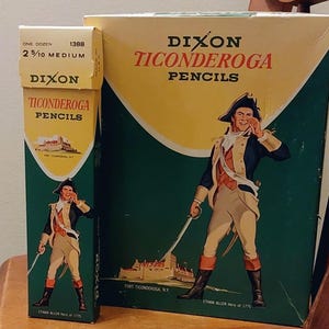 Pode incluir: Duas caixas de lápis Dixon Ticonderoga. A caixa maior apresenta um fundo verde com uma parte superior amarela e um desenho de um homem em uniforme da Guerra da Independência. A caixa menor tem o mesmo design. O texto "Dixon Ticonderoga Pencils" é visível.