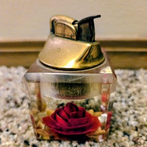 May include: A square, clear acrylic table lighter featuring a brass-toned metal top mechanism. Encased within the transparent base is a single, deep red rose blossom with visible green leaves, creating a floral display inside the lighter body.