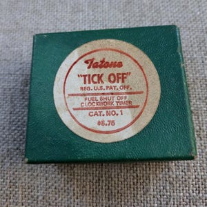May include: A square green box featuring a circular white label with red text that reads Tatone TICK OFF REG. U.S. PAT. OFF. FUEL SHUT OFF CLOCKWORK TIMER CAT. NO. 1 $5.75. The box has a textured surface and is presented from a top-down perspective.