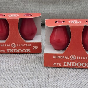 Könnte beinhalten: Zwei rote General Electric C7 1/2 Innenraum-Glühbirnen in ihrer originalen roten Pappverpackung. Jede Schachtel enthält zwei Glühbirnen und zeigt das GE-Logo, den Text General Electric, C7 1/2 INDOOR und eine Preisangabe von 25 Cent in Weiß.