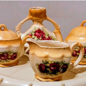 May include: A vintage tea set in cream and peach tones. The set comprises a teapot, sugar bowl, creamer, and a covered bowl. Each item is adorned with red roses and green foliage.