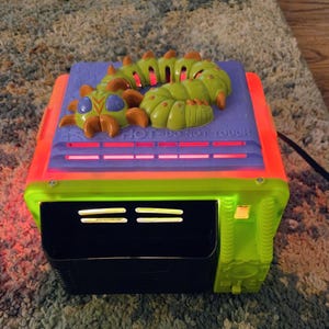 Puede incluir: Un horno de juguete de plástico verde lima y morado con una figura de oruga verde encima. La superficie morada presenta el texto en relieve HOT DO NOT TOUCH. El frente incluye una puerta de horno negra, un botón amarillo y luz naranja.