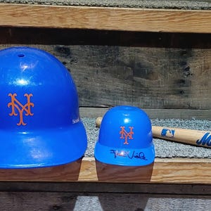 May include: A blue baseball helmet and a smaller blue helmet, both with the orange and blue NY Mets logo. A wooden baseball bat with the Mets logo is also present. The smaller helmet has a signature on it.